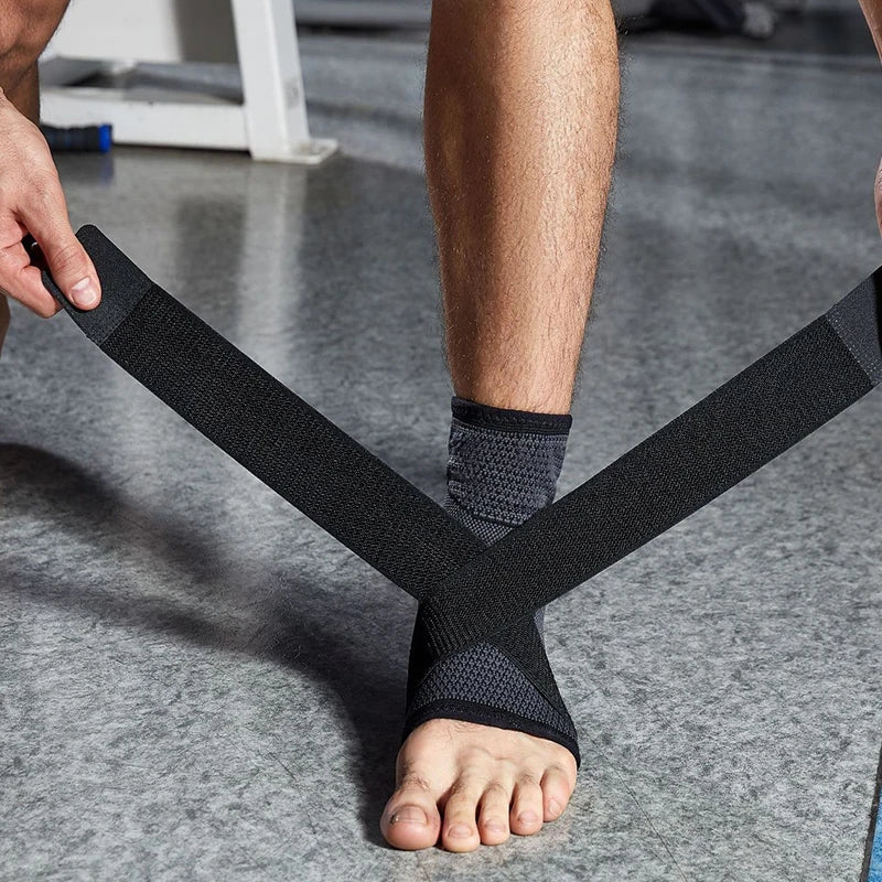 Nahaufnahme einer Person im Fitnessstudio, die eine dunkelgraue Knöchelbandage anlegt. Die Hände ziehen zwei elastische, schwarze Klettverschluss-Riemen über Kreuz über den Spann und um den Knöchel, um den Fuß zusätzlich zu stabilisieren. Der Hintergrund zeigt einen grauen Studioboden und Fitnessgeräte.